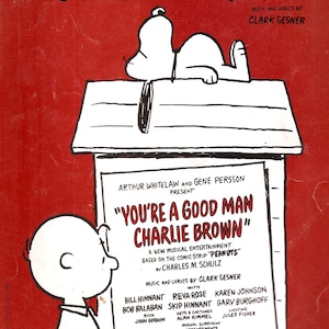 Puede incluir: Un cartel rojo y blanco para el musical "You're a Good Man, Charlie Brown." El cartel presenta una imagen de dibujos animados de Charlie Brown y Snoopy. El musical está basado en la tira cómica "Peanuts" de Charles M. Schulz.