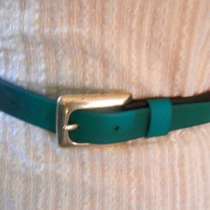 Può includere: Una cintura turchese con fibbia color argento. La cintura è indossata sopra un vestito bianco strutturato. La cintura ha un lato inferiore marrone scuro e una parte superiore turchese. La fibbia è quadrata con bordi arrotondati.