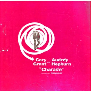 May include: A pink and white movie poster for the film "Charade" starring Cary Grant and Audrey Hepburn. The poster features a white spiral design with an arrow pointing to the right. The text "Charade" is in white letters on a pink background.