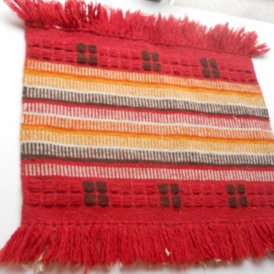 May include: A red woven rug with a fringe border. The rug has a striped pattern in shades of red, orange, yellow, brown, and white. The rug has a geometric design in the center.