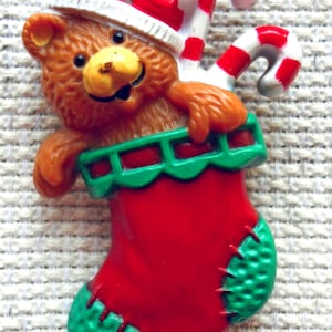 Könnte beinhalten: Ein brauner Teddybär mit einem rot-weißen Weihnachtsmann-Hut schaut aus einem roten Strumpf mit grünem Besatz. Der Strumpf hat einen Zuckerstock, der aus der Oberseite herausragt.