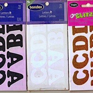 May include: Three packages of iron-on letters. The first package is black Bondex letters, the second is white Bondex letters, and the third is gold glitter Wrights Glitzerz letters. Each package contains 40 individual letter pieces.
