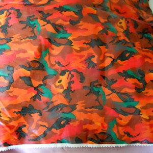 May include: A fabric featuring a camouflage pattern in vibrant shades of orange, red, and black, with accents of bright green. The material has a smooth, slightly reflective surface and a zigzag-cut edge along the bottom of the textile piece.