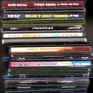 May include: A stack of compact discs with various album titles and artists visible. Titles include "Hits of Stevie Wonder", "Joe Dassin Les Champs Elysees", and "Billboard Top 10 Karaoke". The CDs are in clear plastic cases.