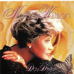 May include: A woman with short, gray hair is looking off to the side. She is wearing a black lace top and has a butterfly on her shoulder. The text "Day Dream" is written in pink letters below her.