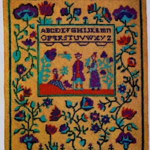 May include: A yellow cross-stitch sampler with a colorful floral border. The center features a scene of three figures in a landscape with a dog. The alphabet is stitched above the scene.