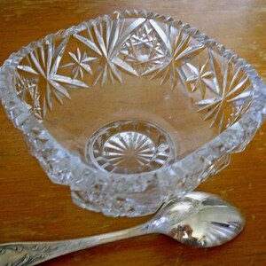 May include: A clear glass bowl with a starburst pattern. The bowl is sitting on a wooden surface with a silver spoon next to it.