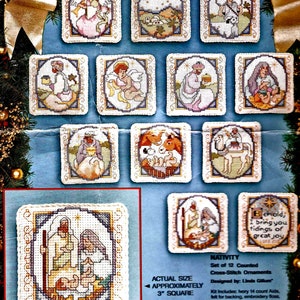 Puede incluir: Un conjunto de 12 patrones de adornos de punto de cruz contados que representan escenas de la Natividad. Cada patrón mide aproximadamente 7,6 cm cuadrados e incluye instrucciones para hilo de bordar, tela Aida, fieltro para el respaldo y una aguja. El texto de la imagen dice "Behold, I bring you tidings of great joy."