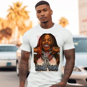 May include: White t-shirt featuring a cartoon illustration of a person with long dreadlocks, a gold chain, and the text "KING VON." The graphic is in shades of brown, gold, and blue. The shirt has a crew neck and short sleeves.