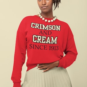 May include: A red sweatshirt with the text "Crimson and Cream Since 1913" in black and white lettering. The model is wearing a white pearl necklace.