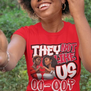 May include: Red t-shirt with a graphic print of two Black women with the text "They Not Like Us 00-007" in white.