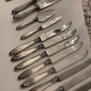May include: An assortment of antique silver cutlery, including knives, a cake server, and a carving fork. The handles feature an ornate floral design. The blades are shiny and reflect the light. A vintage collection for serving.
