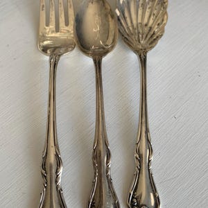May include: Three antique silver serving utensils: a fork, a spoon, and a shell-shaped spoon. The fork has four tines, and the spoon has a rounded bowl. The shell-shaped spoon has a detailed, fan-like design. All pieces have ornate handles.
