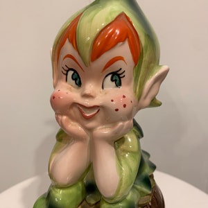 May include: A ceramic figurine of a green elf with red hair sitting on a brown barrel. The elf is wearing a green hat and has a mischievous expression on its face.