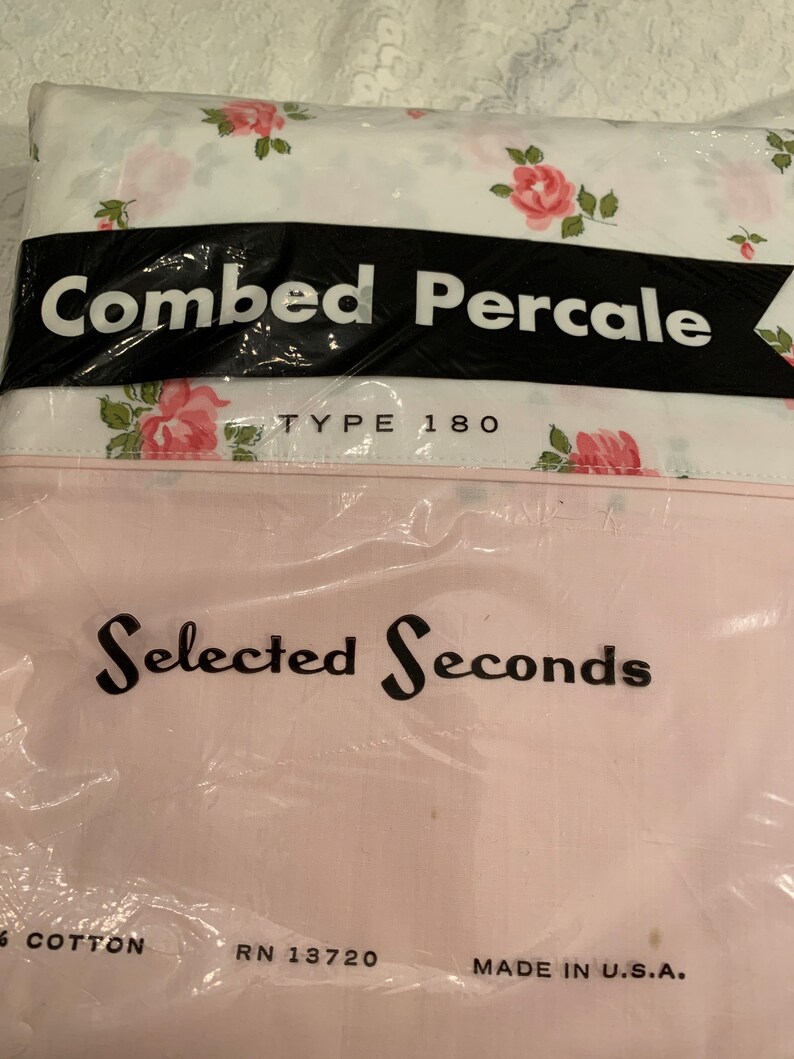 May include: A white sheet with a pink floral pattern, packaged in plastic. The packaging has black text that reads "Combed Percale Type 180" and "Selected Seconds". The bottom of the packaging has black text that reads "6 Cotton RN 13720 Made in U.S.A."