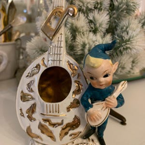 May include: A vintage ceramic figurine featuring a white mandolin with gold accents and a small elf playing a miniature mandolin. The elf wears a blue outfit and a pointed hat. The figurine has a decorative base.