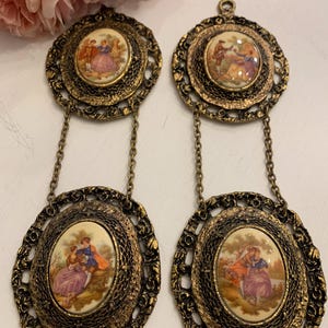 May include: Pair of vintage gold-toned earrings. Each earring features a round top and an oval bottom, connected by a chain. The centers display a painted scene of people in period clothing. The earrings have an ornate filigree design.