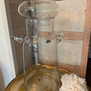 May include: A clear glass jar with a round lid and two handles. The jar is sitting on a gold tray with a white flower.