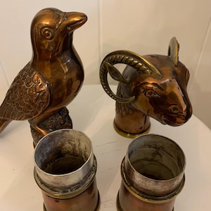 May include: A collection of copper and brass decorative items. Includes a bird figurine, a ram head, and two cylindrical containers with silver rims. The items are arranged on a white surface, showcasing their metallic sheen and intricate details.
