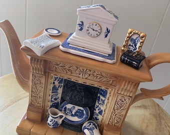 May include: A ceramic teapot shaped like a fireplace with blue and white details. The teapot has a clock, a book, and a small plate on top. There are two small cups and a plate inside the fireplace.
