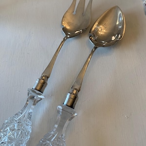 May include: A silver salad fork and spoon set. The utensils have clear, faceted crystal handles and silver metal tops. The fork has three tines, and the spoon has a rounded bowl. The set is designed for serving salads.