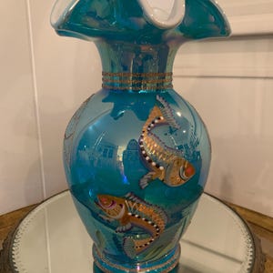 May include: A turquoise iridescent glass vase with a ruffled top and gold trim. The vase is decorated with two painted fish in shades of gold, brown, and white.