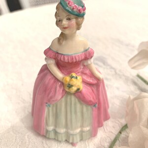 May include: A porcelain figurine of a woman wearing a pink dress, a green hat with pink flowers, and holding a bouquet of yellow flowers.