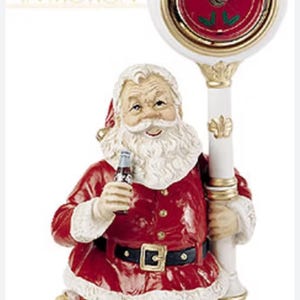 May include: A Santa Claus figurine holding a Coca-Cola bottle, with a clock featuring the Coca-Cola logo. The clock is white with gold accents. Santa wears a red suit with white trim and a black belt. A sack of toys sits at his feet.