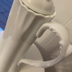 May include: A white porcelain coffee pot with a lid. The pot has a decorative handle and a scalloped edge. The lid has a swan-shaped handle. The pot is sitting on a white surface.
