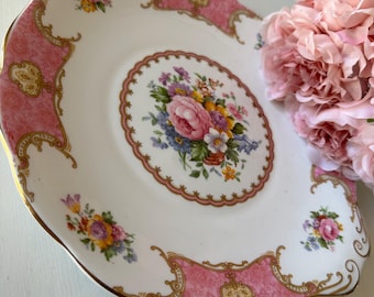Puede incluir: Un plato de servicio de porcelana blanca con borde festoneado y ribete dorado. El plato presenta un ramo floral central de flores rosas, amarillas y azules, rodeado por un borde rosa con volutas doradas y detalles florales adicionales.