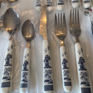 May include: A collection of stainless steel silverware with white handles featuring a blue and white pagoda design. The set includes spoons, forks, and knives, all individually wrapped in clear plastic.