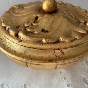 May include: A gold-colored, ornate trinket box with a round shape and a decorative lid. The lid features a detailed, embossed design with a small, spherical knob on top. The box has a distressed, antique appearance.
