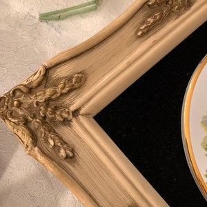 以下が含まれることがあります： 金色のアクセントが付いた白い装飾的なフレームが、花柄の磁器のプレートを囲んでいます。フレームは使い古された感じで、ビンテージな外観をしています。