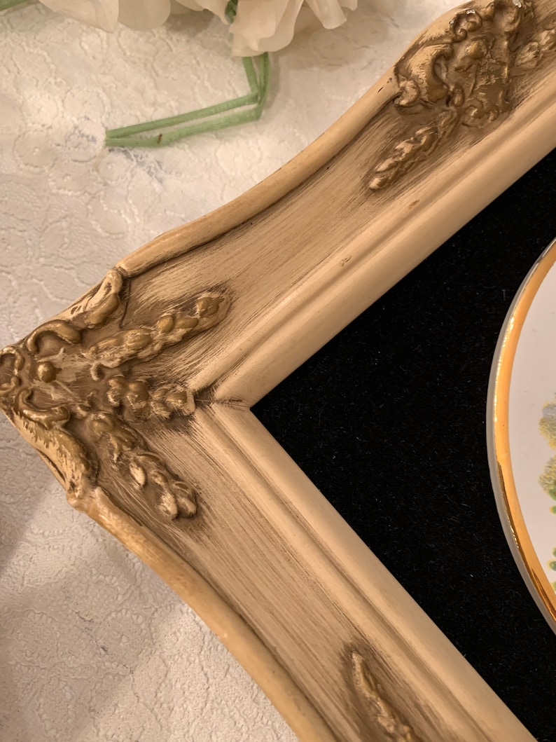 以下が含まれることがあります： 金色のアクセントが付いた白い装飾的なフレームが、花柄の磁器のプレートを囲んでいます。フレームは使い古された感じで、ビンテージな外観をしています。