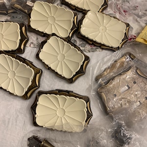 May include: A collection of antique brass and white ceramic wall plates with a floral design. The plates are in various sizes and shapes, and some have a raised edge. The plates are packaged in plastic bags.