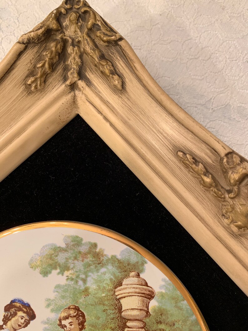 以下が含まれることがあります： 金色の縁取りが施された、額縁に入った磁器製のプレート。プレートには、噴水を背景に庭園にいる2人の女性を描いたシーンが描かれている。フレームは薄いクリーム色で、金色のアクセントが施されている。