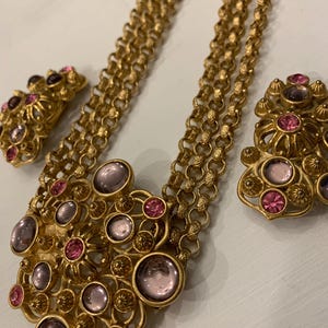 May include: A gold tone necklace with a large pendant featuring a floral design with pink and purple stones. The necklace comes with a pair of matching earrings.
