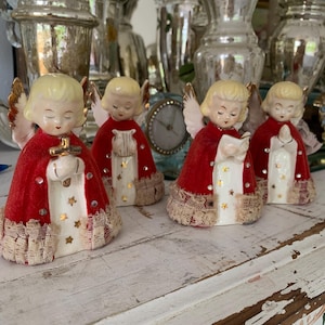 May include: Four vintage ceramic angel figurines with blonde hair, red velvet robes, and gold wings. The angels have closed eyes and are holding various objects. The robes are trimmed with lace and adorned with gold stars. They are arranged on a distressed white wooden surface.