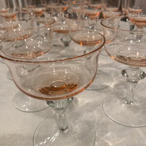 May include: A set of vintage pink glass champagne coupes. The glasses have a delicate pink rim and are arranged in a row on a white tablecloth.