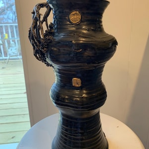 May include: A dark blue ceramic vase with a textured surface and a unique handle resembling intertwined branches. The vase has a bulbous shape with a narrow base and a wide opening. Two gold-colored medallions are affixed to the vase.