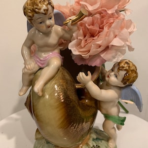 May include: A ceramic vase shaped like a seashell, adorned with two cherubic figures and filled with pink flowers. The cherubs have brown hair, blue wings, and are wearing pink and green accents. The vase has a gold-colored horn.