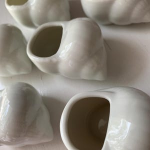 May include: A collection of six small, off-white ceramic seashell-shaped containers. Each shell has a smooth, glossy finish and a hollow interior, perfect for holding small items. The containers are arranged on a white surface.