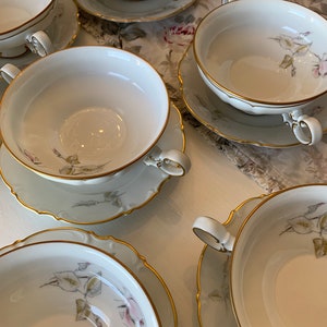 May include: Set of six white porcelain soup bowls with gold trim and floral designs. Each bowl has two handles and comes with a matching saucer.