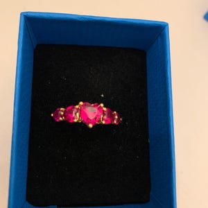 May include: A gold-toned ring with five heart-shaped, bright pink gemstones. The ring is presented in a blue box with a black velvet interior, creating a striking contrast. The gemstones are arranged in a row.