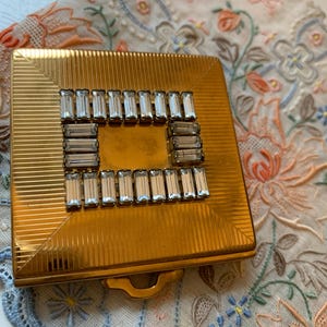 May include: A vintage gold-toned compact with a rectangular shape and a decorative border of clear, rectangular rhinestones. The compact has a ribbed texture and a small handle. The background features a floral embroidered cloth.