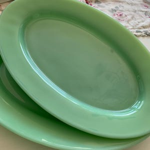 May include: Two oval-shaped plates made of smooth, translucent light green glass. The plates feature a glossy finish and a simple, clean design with a wide rim. The light green color is consistent across both pieces, which are stacked on top of each other.