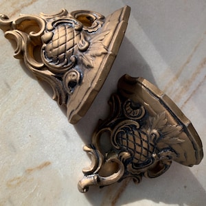 May include: Two ornate gold-painted metal shelf brackets with intricate scrollwork and a pineapple design.
