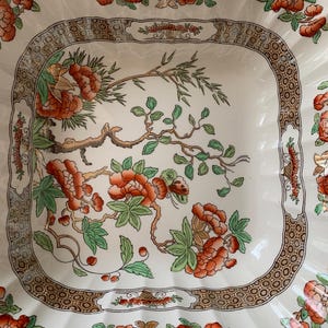 May include: A white ceramic serving dish with a scalloped edge and a square center. The dish features a floral design with orange flowers, green leaves, and brown branches. A brown and gold border frames the floral design.