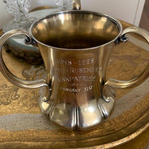 May include: A silver cup with two handles, featuring engraved text: "1948-1955 Otto & Rosemary Kirkpatrick January 31st." The cup has a flared body and sits on a decorative, gold-toned tray. A glass candle holder is visible in the background.