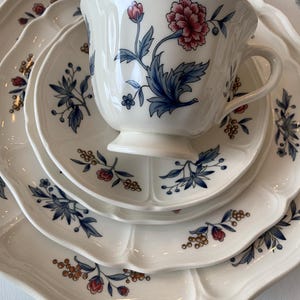 May include: A set of four white ceramic plates with a blue and red floral pattern. The plates have a scalloped edge and are stacked on top of each other. A white ceramic teacup with a blue and red floral pattern is on top of the plates.